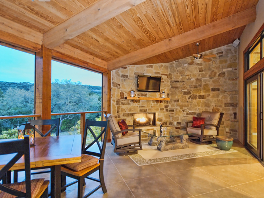 Covered patio with fireplace overlooking the Texas hill country outside of Wimberley. TV mounted on a stone wall. Custom built home by SB Thompson Construction and Contractors.
